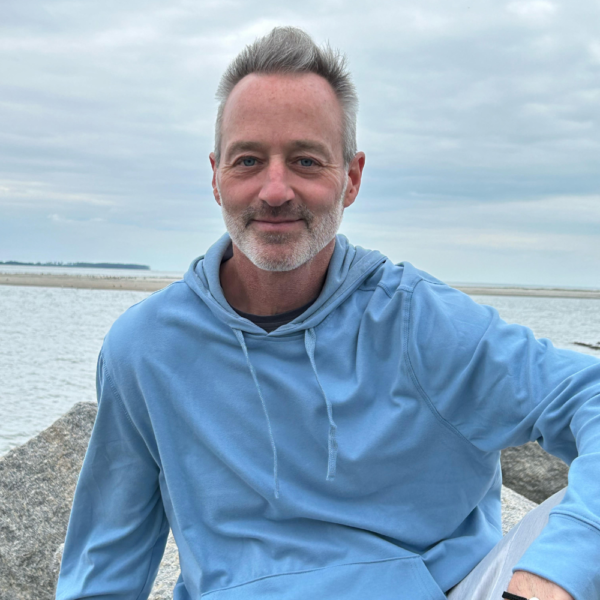 Jim DeBetta in a blue hoodie sitting on rocks by the water.