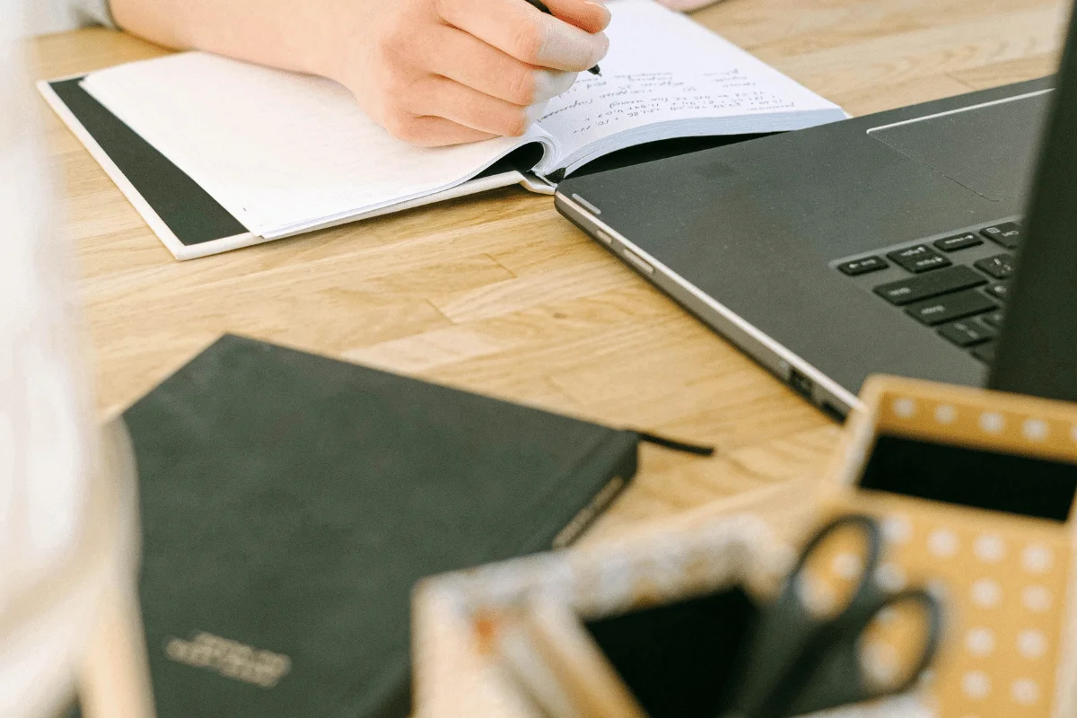 A close-up of a person writing in a notebook beside a laptop on a wooden desk.