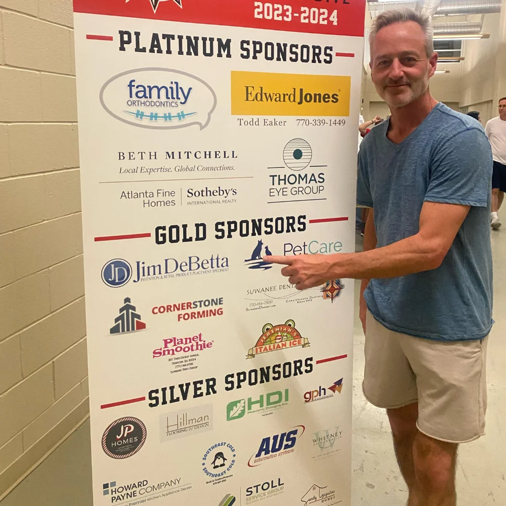 Jim DeBetta points at a sponsor banner for an event categorizing sponsors by platinum, gold, and silver.